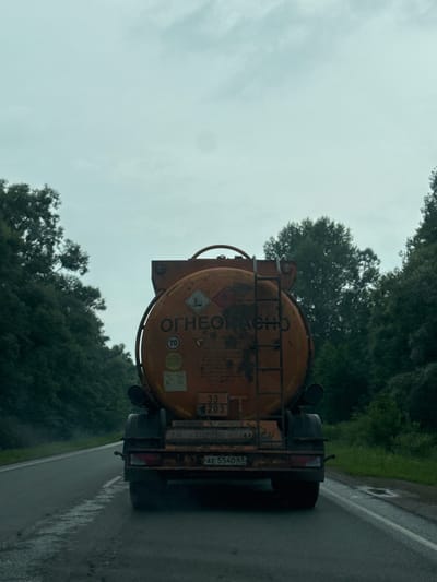 The image shows the rear view of an orange tanker truck on a wet, grey road lined with dense green trees on both sides. The sky is overcast with a pale, cloudy hue. The truck has markings on its tank, including hazard symbols and Russian text that appears to read "ОГНЕОПАСНО" (flammable). There is also a license plate on the back of the truck that reads "AE 5540 67". The truck is moving away from the viewer, down the road. The overall impression is one of a damp, overcast day on a rural or semi-rural road in Russia, with the truck appearing to be transporting hazardous materials. No people are visible in the frame. The location is confirmed as Irnyksh, Russia, based on the license plate format which corresponds to Russian plates.