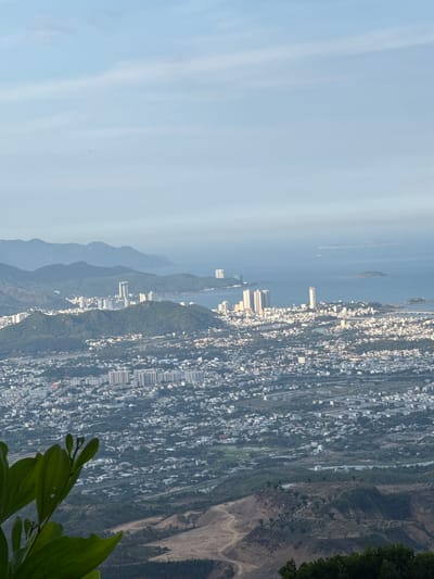 The image captures a panoramic view of the city of Diên Khánh, Vietnam, as seen from a high vantage point, likely a mountain overlooking the urban landscape. In the foreground, a cluster of vibrant green leaves and stems of a plant partially obscures the view, adding a natural element to the scene. The middle ground is dominated by a sprawling urban area characterized by a dense network of buildings and roads, with a mix of low-rise residential structures and several taller, modern buildings, including some high-rises, clustered together. The city extends towards the sea, which forms the background. Distant, hazy mountains rise behind the city, and several small islands are visible in the calm, blue ocean. The sky is a pale blue with faint, wispy clouds, suggesting it is either early morning or late afternoon. The overall impression is one of a coastal city nestled amongst rolling hills and overlooking a serene body of water.