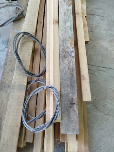 This is a top-down, medium shot of a pile of wooden planks and two coiled lengths of metal wire. The planks are of varying widths and ages; some are weathered and grey, while others appear newer and lighter in color. They are stacked on a concrete or dirt surface. The wire is thick and appears to be made of multiple strands twisted together, forming two distinct loops. One coil is positioned above the other. The scene is outdoors, with natural light suggesting daytime. There are no people or animals visible. The setting appears to be a workshop, construction site, or storage area for building materials. There is no visible text in the image. The overall impression is one of raw materials and a practical, utilitarian setting. The location context of Крывасельскі сельскі Савет, Belarus, is not directly evident from the image itself but provides an external reference point for the geographical location.
