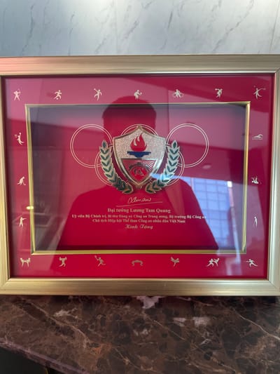 The image shows a framed award with a red background and gold trim. The award is displayed on a marble surface. The frame is made of gold-colored metal and contains small, gold silhouettes of various athletes around the border. In the center of the award, there is a crest with a torch and laurel leaves. Below the crest, there is text in Vietnamese. The text identifies the recipient as General Luong Tam Quang, a member of the Central Committee of the Communist Party of Vietnam, Deputy Minister of Public Security, and Chairman of the Vietnam Sports Association for Public Security Personnel. The award is presented to him for his contributions. The background of the award also shows a reflection of a building with a grid-like window pattern, suggesting the award might be located in an indoor setting within a city. While the exact location in Thủ Đức, Vietnam is not specified, the presence of a modern building reflected in the award suggests an urban environment, possibly an office or a ceremonial hall.