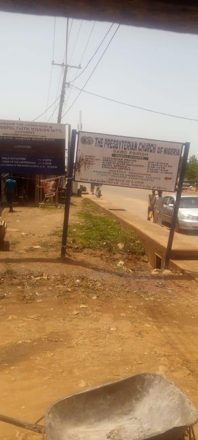 The image depicts a dusty roadside scene in Yelwa, Nigeria. Two church signs dominate the view: one for "Gospel Faith Mission Int'l" and another for "The Presbyterian Church of Nigeria, Sabo Parish," detailing weekly activities and service times. A few people are visible, including one man walking near the Gospel Faith Mission sign, and two people standing beside a silver car parked on the street near the Presbyterian Church sign. Overhead power lines are also apparent. A wheelbarrow is present in the foreground. The environment appears arid with exposed dirt and sparse vegetation.
