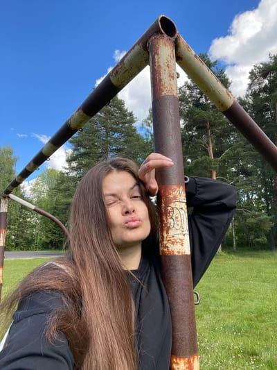 A young woman is lying down outdoors, resting her head on a rusty metal bar. She is wearing a black hoodie. Her eyes are closed, and her lips are puckered, as if she is about to blow a kiss or make a kissy face. Her long, brown hair is spread out around her head.
The setting appears to be a grassy field with trees in the background, suggesting a park or a rural area. The sky is blue with some white clouds, indicating it is daytime and likely pleasant weather. The metal bar she is lying on looks like part of an old piece of playground equipment or sports equipment, such as a goalpost or a swing set. It is weathered and shows signs of rust.
There is some text visible on the metal bar, but it is difficult to read clearly. It appears to be handwritten in black marker.
The overall mood of the image is relaxed and playful, with the woman's expression and pose conveying a sense of lightheartedness. The orientation of the image is rotated, making it appear sideways.