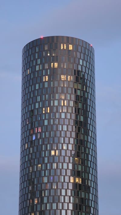 The image shows the Beetham Tower, a skyscraper in Manchester, United Kingdom. The tower is cylindrical and covered in glass windows, some of which are lit from within. Two red lights are visible at the very top of the building, likely aviation warning lights. The sky behind the tower is a pale blue, suggesting it is either dusk or dawn. The overall impression is of a modern, imposing structure set against a calm, atmospheric sky.