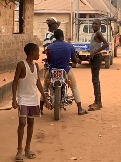 The scene depicts a daylight street view in Aguleri, Nigeria, on a reddish-brown dirt road. In the foreground left, a young male with short dark hair wears a white tank top, purple athletic shorts with a yellow stripe, and light-colored slip-on sandals, looking towards the right. Directly behind him, an adult male is seated on a motorcycle, facing away from the viewer. He wears a blue t-shirt with red accents on the shoulders, light brown trousers, and dark slip-on sandals. The motorcycle features a chrome exhaust, red taillight assembly with two circular lights (appearing as "88"), and a visible Nigerian license plate.

To the left of the motorcycle, partially obscured by the rider, an adult male stands wearing a blue and white striped long-sleeve shirt and a light-colored baseball cap, looking towards the right. To the right of the motorcycle, a young male stands with his hands on his hips, wearing a grey tank top, dark jeans, and patterned brown slip-on sandals. He is looking directly at the camera with a slight smile.

In the background, a white pickup truck with a blue "Panasonic" decal on its tailgate is visible, featuring a metal frame rack above its bed and wooden side railings. Further background elements include faded block structures, one with an open window or missing section, and corrugated metal roofs under bright natural lighting. Scattered debris, including small leaves and pieces of wood, is present on the dirt road.