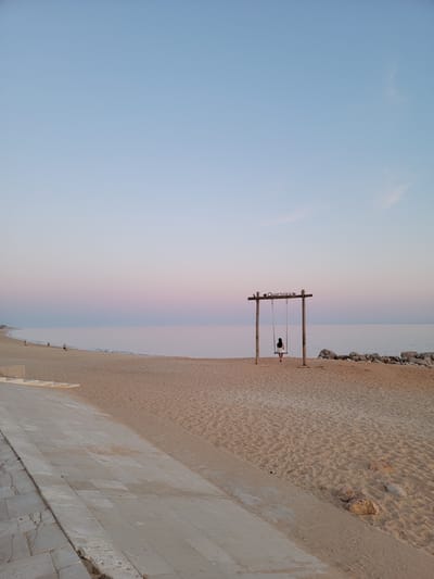 The image shows a beach in Quarteira, Portugal at dusk. The sky is a gradient of pale blue to pink. The sea is calm and meets the sandy beach. On the beach, a wooden swing set with "Quarteira" written across the top beam is prominent. A person, dressed in white, is sitting on the swing. To the left, a paved walkway with steps leads down to the sand. Further down the beach, a few other people are visible walking along the shoreline. Rocks are visible near the edge of the water to the right of the swing. The sand is imprinted with ripples and small footprints.