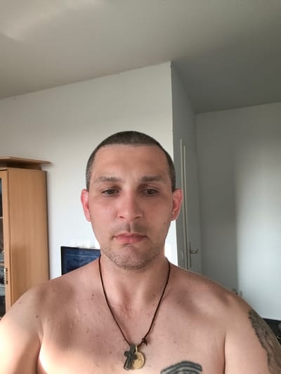 A man is captured in a front-facing, medium shot, shirtless and looking directly at the camera. He has short, dark hair and a light stubble on his face. He is wearing a dark necklace with a pendant that appears to be a cross. His skin has some freckles and what seem to be minor scars on his chest. A tattoo is partially visible on his right shoulder.

In the background, to the left, a light-colored wooden cabinet with glass doors is visible. Behind him, a television screen displays some text, possibly subtitles or a menu, in what appears to be a Cyrillic script. Further to the right, a white door with a silver handle stands slightly ajar. The walls are painted a neutral, light color. The lighting suggests it is daytime. The overall setting appears to be an indoor residential space. The man's expression is neutral, and he does not appear to be actively engaged in any specific activity other than posing for the photo.