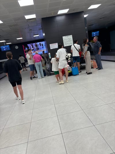 In the city of Malye Kabany, Russia, an airport terminal is depicted with a group of people standing and interacting. The scene is captured from a slightly elevated perspective, looking down and across a tiled floor.
In the foreground, a woman in a black t-shirt and black shorts walks away from the camera, facing left. She has sunglasses on her head and is wearing white sneakers.
To the left, a man with dark hair, wearing a grey t-shirt and blue shorts, is speaking on his phone. Next to him, a woman in a bright pink t-shirt and light blue jeans stands facing away from the camera. Behind them, a man in a white t-shirt and dark shorts also stands.
In the mid-ground, a group of people are gathered. A woman in a white t-shirt and white shorts stands facing right, holding a stuffed panda. Beside her, a young girl in a white t-shirt and green shorts is being spoken to by a woman in a black and white striped top and light-colored pants. A blue suitcase is on the floor near them. Further to the right, a man in a striped blue and white shirt and grey pants is talking to another man in a white t-shirt and dark shorts. Several bags are visible near this group.
The environment appears to be an indoor space with a high ceiling featuring multiple fluorescent lights. A large screen displaying flight information or advertisements is visible on the left wall. A clock is also mounted on the wall. The walls are dark and modern, with some signage. The floor is tiled with large, light-colored square tiles. The overall impression is of a busy airport waiting area.