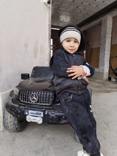 A young child stands leaning against a black ride-on toy SUV in an outdoor, covered area in Limbur, Uzbekistan. The child, appearing to be a toddler or preschooler, has light skin, dark eyes, and a slight smile, looking directly at the viewer. They are dressed in a beige and black striped knit beanie, a dark hooded jacket, and dark sweatpants with a white Adidas logo on the left thigh. The child's arms are crossed over their chest; a small ring is visible on the right ring finger, and the left sleeve cuff is white with a red pattern. The toy SUV is black, features a Mercedes-Benz star logo on its vertical-slat grille, and has a "SPEED 800" plate and silver winch replica on its dusty front bumper. The ground is a light grey concrete surface. To the left, a plain, light-colored stucco wall with minor discoloration and a white electrical box is visible. To the right and behind the child, the environment includes a tiled ceiling with a grid pattern, two decorative concrete pillars with patterned finishes, and a dark opening leading further into a structure, with a partial view of a dark bicycle frame on the far right.