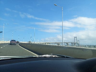The image captures a bright daytime view from the interior of a vehicle, looking forward along an elevated highway or bridge. The lower part of the frame shows the car's dashboard and the base of the windshield, including a wiper blade.

The highway features at least two lanes of traffic, clearly marked with white lines. Numerous modern streetlights on tall poles line both sides of the roadway, extending into the distance. Concrete barriers or guardrails separate the main highway from a parallel, lower structure on the right. Several vehicles are visible ahead on the road, including a dark pickup truck with a light bar on its roof.

In the mid-ground and background, a prominent cable-stayed or suspension bridge spans the horizon, characterized by its large A-frame pylons and visible cables. This iconic structure is identifiable as the Maputo-Katembe Bridge in Maputo, Mozambique, aligning with the provided location context. The sky above is a vibrant blue with scattered white, wispy clouds, indicating clear and sunny weather conditions. No visible text or discernible human activity beyond driving is present. The overall scene suggests a modern urban or coastal infrastructure setting.