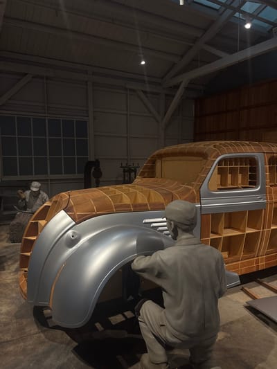 This image depicts a diorama in the Toyota Commemorative Museum of Industry and Technology in Nagoya, Japan, illustrating the automobile manufacturing process. In the foreground, a gray, life-sized mannequin of a man is kneeling and appears to be working on the fender of a car body. The car body itself is under construction, with a wooden frame visible beneath a partially completed silver exterior. Further in the background, another mannequin of a man in a cap is seated and holding a tool, suggesting assembly or shaping work. The environment is an indoor workshop setting, with a concrete floor, corrugated metal walls, and a wooden beamed ceiling with exposed lighting. Large windows with a grid pattern are visible on the back wall, and industrial machinery is present in the mid-ground. The lighting is dim, with spotlights illuminating the main subjects.