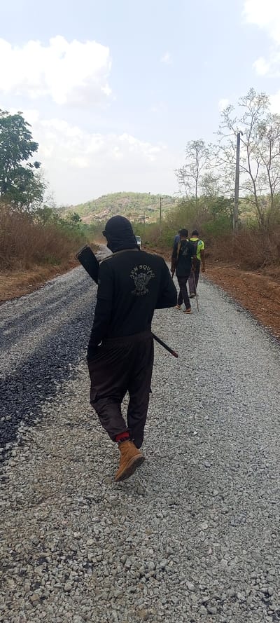 A group of four or more individuals is captured from behind, walking away from the camera on a gravel road under a bright, partly cloudy sky. The setting appears to be rural or semi-rural, characterized by dry, sparse vegetation and a hilly landscape in the background.
The closest individual, central in the frame, wears a black long-sleeved shirt, dark pants (possibly cargo pants), and tan/brown work boots. A red and black checkered pattern is visible on their socks above the boots. Their head is covered by a black balaclava or similar headwear, obscuring their face. On the back of their shirt, a distinct white logo is visible, featuring an emblem that appears to be a winged creature or skull with crossed weapons, above which is the text "ARM SQUAD" and below it, "SLAYERS." This person carries a dark, elongated object resembling a rifle or a long tool over their left shoulder, and another thin, dark object, possibly a sheathed blade or stick, is tucked into their waistband on the right side.
Further down the gravel road, two other individuals are visible. The one directly in front also wears a dark shirt with what appears to be a similar logo on the back, though less clearly discernible. The person further back wears a bright greenish-yellow shirt. All individuals are walking in the same direction.
The road itself is composed primarily of light grey gravel. On the left side of the road, there's a darker, possibly asphalted or freshly laid section of road material, suggesting construction or repair work. In the far distance, a light-colored vehicle, possibly a van or SUV, is faintly visible, stirring up a small dust cloud. The background features several sparse trees and bushes, indicative of a dry environment, with a prominent hill or small mountain range dominating the horizon. A utility pole stands on the right side of the road in the mid-distance. The bright daylight and clear sky suggest it is daytime, likely during a warm period.