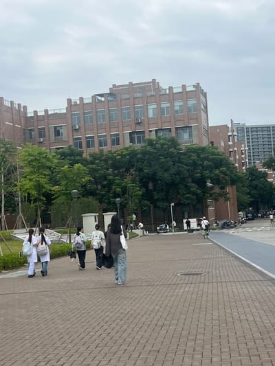 This is a medium-shot, eye-level, outdoor photograph taken on a cloudy day in the Liandu District of China. The scene depicts a paved walkway in front of a large, multi-story brick building with numerous windows and a flat roof. The building is surrounded by lush green trees. Several individuals are walking away from the camera on the walkway. Two individuals in the foreground are wearing white tops and jeans, each carrying a backpack. Behind them, another person in a light-colored top and jeans is also carrying a backpack. Further back, a person in dark clothing is walking with a white bag. In the background, there are more people, some on bicycles and scooters, and additional buildings. The walkway is made of interlocking bricks, and there is a blue lane on the right side of the frame, possibly a bike or pedestrian path.