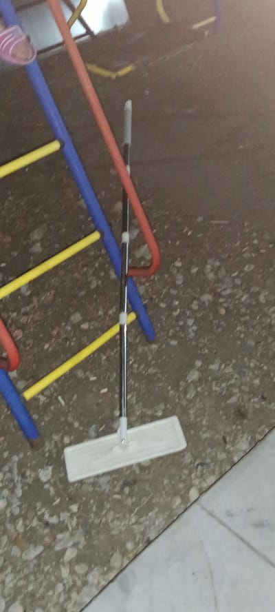 The image shows a mop with a white flat head and a metal handle leaning against a playground structure. The playground structure consists of blue and red pipes, with yellow horizontal bars. A pink sandal is visible on the top left portion of the structure. The ground is covered with small stones and dirt. The scene appears to be outdoors, possibly in a park or courtyard. The lighting suggests it is daytime. There is no specific indication of the location being in Novotitarovskaya, Russia, other than the prompt's assertion.
