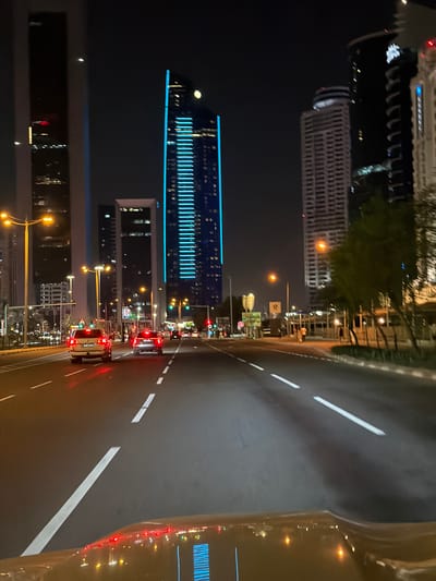 The image displays a vibrant nighttime urban scene in Doha, Qatar, captured from the perspective of someone inside a vehicle. The road, featuring clear white dashed and solid lane markings, occupies the right side of the frame and recedes into the distance. Several cars are visible on the road, with their red taillights illuminating the asphalt, indicating active traffic. A yellow vehicle, likely a taxi, is prominent in the foreground, its brake lights glowing brightly.
On the left side of the frame, a modern cityscape rises against the dark night sky. A striking skyscraper in the mid-ground stands out with its distinctive bright blue vertical and horizontal strip lighting, creating a prominent visual feature. Other tall buildings with illuminated windows are visible in the background and foreground along the street. The area is well-lit by numerous streetlights emitting a warm orange glow, which also illuminates some sparse trees and vegetation situated between the road and the buildings.
Red traffic lights are discernible in the distance, indicating intersections or congestion points. Though some road signs are present, any text on them is unreadable due to distance and image resolution. The overall atmosphere is that of a bustling yet orderly city after dark. The weather appears clear, with no visible precipitation or fog. No clearly discernible people are visible, though a small, distant red figure near a building could potentially be a person.