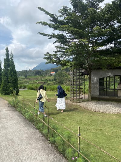 Two people stand in a green yard with tall grass. One wears a yellow top and jeans, while the other wears a dark blue and white outfit. A tree with dense foliage is at the top left. A stone-walled building with a window and a gravel path are behind the people. A metal fence-like structure is near the tree. A mountain and rice paddies are visible in the background under a cloudy sky. A cement path runs along the bottom of the image.