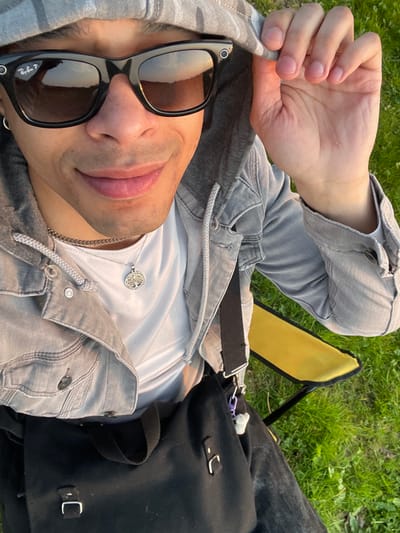 A male subject is depicted seated outdoors on a patch of green grass in Plaisir, France, during what appears to be golden hour. The subject is looking upwards directly at the camera with a slight smile. He wears black Ray-Ban Wayfarer-style smart sunglasses, reflecting a sunset or sunrise horizon. He has a silver hoop earring in his left ear. He is dressed in a white t-shirt, a silver chain necklace with a circular "Tree of Life" pendant, and a grey hooded denim jacket with a lighter grey hood, the hood of which he is adjusting with his right hand. Dark trousers are partially visible. A black shoulder bag with silver buckles and a white object attached by a purple clip rests on his lap or beside him. A yellow folding chair is partially visible on the right side of the frame, underneath the subject's right arm.