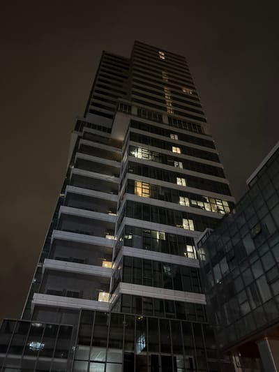 This is a low-angle, full shot of a modern high-rise building at night, located in the Huangpu District of China. The building is tall and slender, with a dark, clouded sky as the backdrop. The facade of the building is a combination of dark glass and white horizontal accents. Many of the windows are illuminated from within, casting a warm glow on the exterior. Some balconies are visible with dark grates. To the right of the main building, a smaller, adjacent structure with a glass facade is partially visible. The overall impression is one of urban nocturnal architecture.