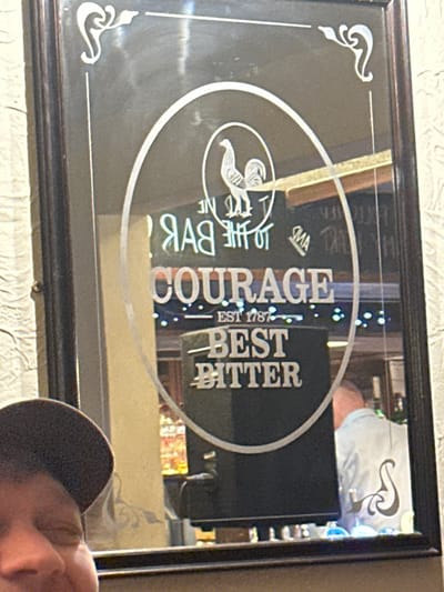 A framed mirror displays an advertisement for Courage Best Bitter, established in 1787. The image includes a logo of a horse inside a circle, with text reading "At the bar, Courage Best Bitter." The mirror reflects the surrounding environment, including a person wearing a hat in the corner. The image is slightly rotated.