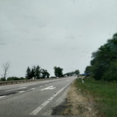 The image depicts an asphalt road with white lane markings, viewed from a slightly elevated perspective. Trees and grass line the right side of the road. The sky is overcast, suggesting a cloudy or overcast day. The image appears to be taken from a moving vehicle, as indicated by the motion blur and the angle of the shot. There are no people or specific objects readily identifiable, beyond the road and surrounding vegetation. The scene is a rural or semi-rural road, possibly a highway or main road. No text is visible in the image. The overall impression is of a typical roadside view on a day with subdued lighting.