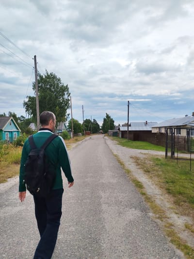 In the city of Shalsky, Russia, a man with a black backpack walks away from the camera on an asphalt road. He is wearing a green jacket with white stripes on the sleeves and dark pants. The road is lined with houses and trees on both sides. The sky is cloudy. The scene appears to be a rural or semi-rural setting.