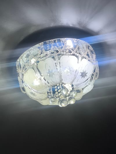 A close-up, low-angle shot captures a decorative ceiling light fixture. The fixture appears to be made of glass and crystal elements, with a spherical shape. It features frosted glass panels with etched floral patterns and is adorned with multiple strands of small, clear beads and larger, faceted crystal drops. The light source within the fixture is visible, emitting a bright, diffused light. Shadows and light refractions are cast onto the dark ceiling, creating a textured, ethereal effect. The image is undated and location-unspecified, therefore it cannot be definitively placed within the city of Kokand, Uzbekistan.
