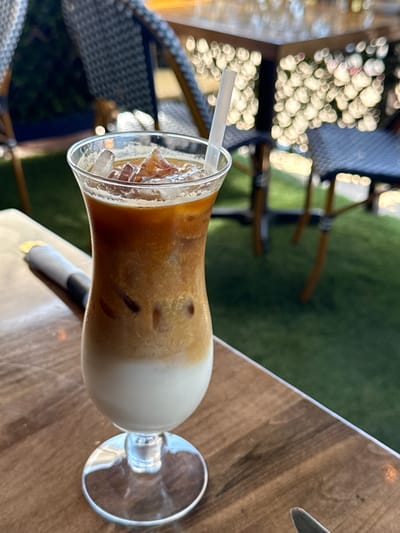 The image shows a close-up of a glass of iced coffee on a wooden table, with a straw in the glass. The background includes outdoor seating with chairs and a blurred green area. The location is Chula Vista, United States of America.
