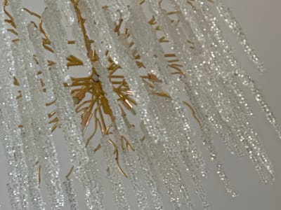 The media file shows a close-up, abstract view of a decorative chandelier. The chandelier features multiple strands of clear, possibly glass or crystal, beads that hang downwards. These strands are attached to a central, ornate fixture with thin, golden-colored metallic elements that branch out like delicate twigs or icicles. The lighting in the scene is bright, causing the beads to reflect and sparkle, creating a glittering effect. The background is a plain, light-colored surface, likely a ceiling, which is out of focus. The image is slightly blurred, giving it a soft, dreamy quality. There is no visible text, people, or clear indication of time of day or weather. The focus is solely on the intricate details and sparkling texture of the chandelier itself. The setting appears to be indoors, in a well-lit space.