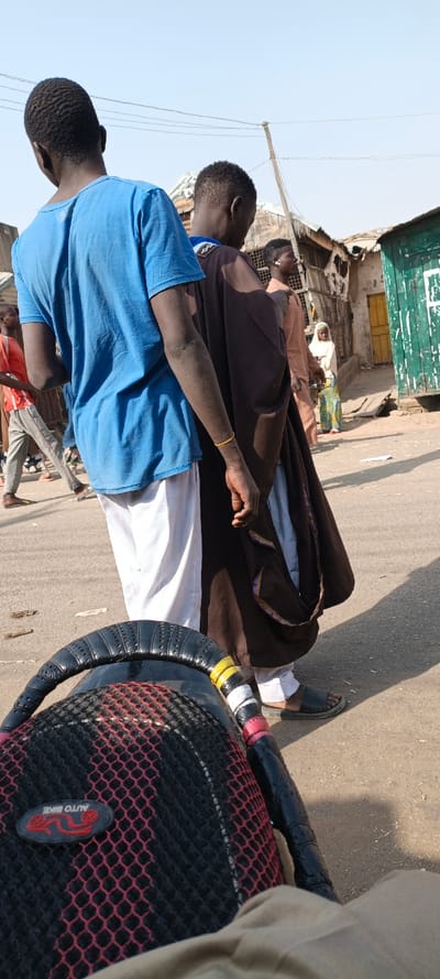 The image captures a street scene in Kaura Goje, Nigeria, from the perspective of someone seated on a motorcycle. In the immediate foreground, the black seat and grab rail of a motorcycle are visible, featuring a red and black mesh cover with an "AUTO BIKE" patch. The grab rail is wrapped with yellow, white, and red tape.
In the mid-ground, several individuals are present. Closest to the camera, with their back to the viewer, is a dark-skinned male wearing a light blue t-shirt and white trousers. To his right and slightly behind him is another dark-skinned male, also facing away, wearing a dark brown flowing garment over a lighter undergarment and white trousers, with black sandals on his feet. A gold-colored bracelet is visible on his right wrist. Further to the right and partially obscured, a third dark-skinned male is seen in a tan shirt, with an earpiece in his right ear. To the far left, a fourth dark-skinned male in a red t-shirt and grey trousers walks away from the camera. A young female child in a white hijab and a long, colorful patterned dress stands further back on the right side of the street.
The background consists of weathered buildings and structures under a clear, bright blue sky. On the right, a building with a peeling green facade and a yellow door is visible. Another structure behind the child has a lighter wall and a partially open yellow door. Corrugated metal roofs are visible on several buildings. A wooden utility pole stands towards the center-right. The ground is a light-colored asphalt or dirt road, showing distinct shadows cast by the strong daylight.