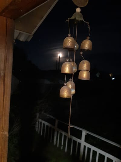 This media file shows a wind chime hanging outdoors at night. The wind chime consists of several bell-shaped elements made of a golden material, possibly metal, with intricate patterns etched onto their surfaces. These bells are suspended from strings, interspersed with small, round beads of a lighter color, possibly pearls or glass. The wind chime is the primary object in the foreground, and its elements are arranged horizontally across the frame. In the background, the scene is dominated by darkness, suggesting it is nighttime. A bright light source, likely the moon, is visible in the sky, casting a soft glow and reflecting on what appears to be a body of water. To the right of the wind chime, a white railing or fence is partially visible, with diagonal slats creating a geometric pattern. To the left, the edge of a building with a sloping roof is visible, constructed with wooden siding. The overall impression is one of a quiet, serene night, possibly in a backyard or on a balcony, with the gentle movement of the wind chime the only implied activity. The location is identified as Shelekhov, Russia. There is no visible text in the image.
