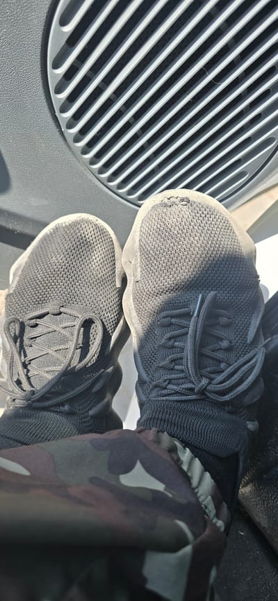 The image shows a pair of dark gray, mesh athletic shoes with black laces, worn by an unseen individual. The shoes are positioned on a gray surface with a ribbed texture, likely a vent or grate, suggesting an indoor environment such as a car interior or building. One shoe exhibits a noticeable tear on the toe. The lower part of the image shows camouflage-patterned fabric, possibly part of clothing. The overall scene lacks any specific indicators of a location within Baku City, Azerbaijan.