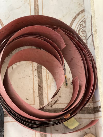 A pile of curved, red metal sheets is coiled on a tiled floor. The tiles are a light beige with brown and darker beige geometric patterns. The metal sheets have a textured surface with a diamond pattern and appear to be used or scrap material. Some debris, including dirt and small pieces of paper, is scattered on the floor and on the metal sheets. A green pen with a yellow cap rests on top of one of the metal sheets. The scene appears to be indoors, possibly in a workshop or storage area, located in Wai A, Nigeria.