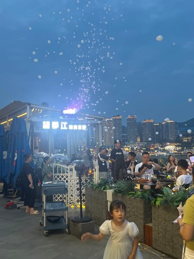 This media file captures an outdoor evening scene, possibly a celebration or gathering, in Jiangbeicheng Street, China. The sky is a deep blue, transitioning from twilight to night, with numerous small, translucent bubbles floating upwards, creating a whimsical atmosphere. A cluster of tall buildings with illuminated windows forms the backdrop, suggesting an urban environment.
In the foreground and mid-ground, several people are visible, some seated at tables, others standing. Many appear to be engaged in conversation or enjoying the event. A young girl in a white dress stands out, looking towards the camera with a neutral expression. The setting includes tables, chairs, and what appears to be a rooftop or elevated patio with some greenery and decorative elements. A sign with Chinese characters is illuminated and mounted on a structure, adding a specific location cue. The presence of umbrellas and what looks like a cart with equipment suggests a planned event. The overall mood seems relaxed and celebratory, enhanced by the soft lighting and the ethereal presence of the bubbles.