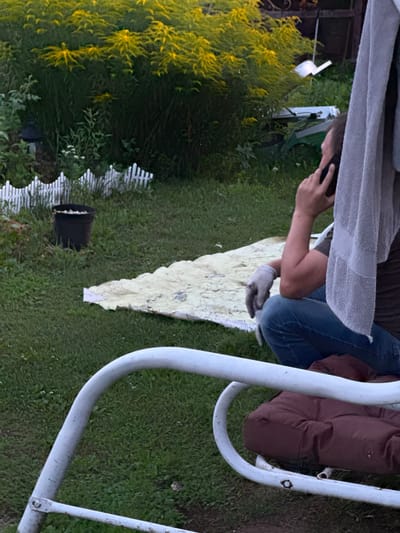 The image depicts an outdoor scene in the city of Belous, Russia, characterized by a garden or backyard setting. In the foreground, a portion of a white metal swing set with a maroon cushion is visible. A person, seen from the side and slightly behind, is seated on the grass, wearing blue jeans, a dark t-shirt, and a grey gardening glove on their left hand. They are holding a dark mobile phone to their right ear, appearing to be engaged in a conversation. A light-colored towel is draped over their shoulder and head. Behind the person, a yellow-flowering plant, possibly goldenrod, dominates the background. Further back, a white picket fence encloses a section of the yard, and a black bucket filled with what appears to be small white objects is placed near the fence. The ground is covered in green grass, with some scattered weeds and small white flowers. The overall impression is of a casual, everyday moment occurring in a residential outdoor space.