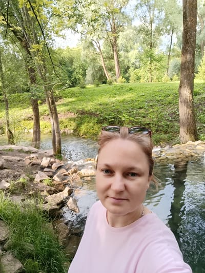 The image is a selfie of a woman in a pink t-shirt standing outdoors near a body of water. She is wearing sunglasses on top of her head and a necklace. The background shows a natural landscape with trees, grass, and rocks. The water appears to be a stream or river. Sunlight is filtering through the trees, suggesting it is daytime. The overall mood of the image is relaxed and natural. The location cues, such as the types of trees and the rocky terrain, are consistent with a natural park or wooded area in Russia, specifically Sdt Shcherbakovka. There is no visible text in the image.