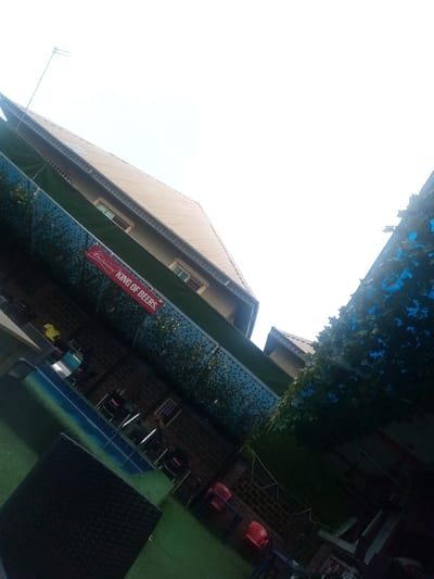 The image shows an outdoor seating area in what appears to be a bar or restaurant. There are tables and chairs arranged on artificial green turf. The area is bordered by a wall decorated with artificial foliage and a "Budweiser KING OF BEERS" sign. Overhead, there are building structures and an antenna visible against a bright sky. The lighting is uneven, with some areas in shadow. The location is Asaba, Nigeria.