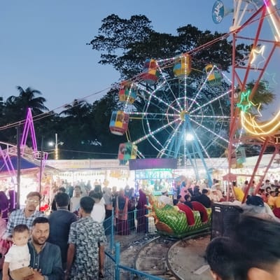 This image captures a vibrant evening fair or carnival scene in Charfasson, Bangladesh, at dusk. The dominant feature is a large Ferris wheel, brightly lit with multi-colored LED lights that illuminate its spokes and colorful gondolas (red, yellow, blue, green). To the right, another fair structure is adorned with glowing neon star and crescent moon shapes, alongside a blue megaphone.

In the mid-ground, a track ride with a distinctive green dragon-shaped car on rails is visible, with several people standing nearby. The scene is bustling with a large crowd of people, including adults and children, many dressed in casual or traditional South Asian attire, moving through the fairground. In the foreground, a man is holding a young child, both looking towards the camera, and another man stands beside them. A blue metal fence separates the onlookers from the ride areas.

The fading blue sky, along with the numerous illuminated rides and stalls (some seen in the background), indicates that the event is taking place as day transitions into night. The atmosphere appears festive and lively, characteristic of a community gathering or exhibition.

Visible text on banners above the crowd, primarily in Bengali, includes phrases such as "জ্য মেলা/প্রদর্শনী-২০২৬ ইং চরফ্যাশন মাসব্যা" (Victory Fair/Exhibition-2026 AD Charfasson monthly/mass-based) and "আয়োজনেঃ চরফ্যাশ" (Organized by: Charfasson). Other fragments like "এসক, ভোলা।" and "সার্বিক সহযোগি" (Overall Collaborator) further indicate a local fair or exhibition, likely in the Bhola district of which Charfasson is a part.