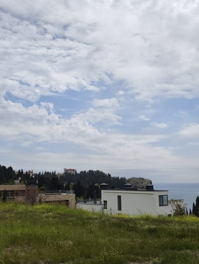 The image shows a landscape scene with a modern white building in the foreground. The building has a flat roof, rectangular windows, and a glass-paneled balcony. To the left, another building with stone cladding is partially visible, surrounded by green grass. Behind the buildings, a hill covered in dark green trees rises, with a large building with a red roof visible at the top. To the right of the hill, a rocky outcrop juts out into the sea, which is visible in the background. The sky is filled with white clouds. The scene suggests a location with a coastal view, possibly a residential area near a forested hillside, with architectural styles and vegetation commonly found in the coastal regions of Ukraine. Given the specification of "Ut'os, Ukraine", this is likely a coastal area, known for its natural beauty and modern architecture. It is not possible to verify people or interactions based on the image.
