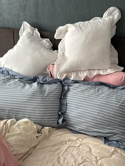 A messy bed with multiple pillows and crumpled sheets is shown in a close-up, eye-level shot. Two large, white, ruffled pillows are stacked at the top, behind a pink bolster pillow. In front of the bolster pillow are two blue and white striped pillows. A light-colored blanket with small dark hearts is draped over the bottom left of the bed, partially covering a pink cloth. The background consists of a dark, textured wall. The scene is indoor, likely a bedroom. There is no indication of Prikubanskii, Russia.