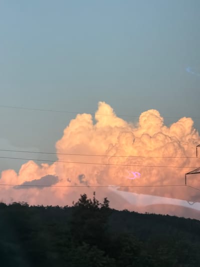 A sky of pale blue and soft pink is dominated by voluminous, illuminated clouds that appear to be catching the light of a setting sun. In the foreground, a dark, hilly landscape is covered with trees, their silhouettes creating a natural horizon line. Several power lines crisscross the frame, adding a man-made element to the scene. A faint, purple, abstract shape is visible in the sky, possibly an artifact of the camera or a light anomaly. The image is a landscape captured from a moving vehicle, as evidenced by the slight blur of the foreground trees. The specific location in Rajana, Bulgaria, is not discernible from the visual content of the image alone.