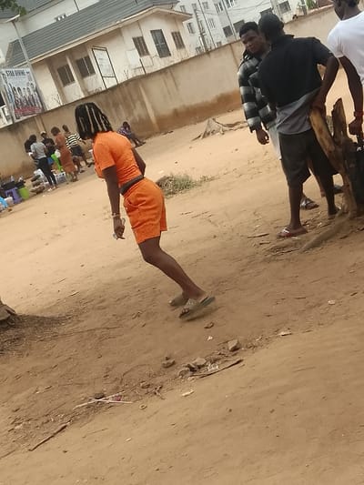 The image displays an outdoor street scene in Umuokpu, Nigeria, primarily characterized by reddish-brown dirt ground. In the foreground, a person with dark skin, appearing female, is depicted from a rear-left perspective, angled slightly forward, as if in motion. This individual wears a matching orange short-sleeved crop top and orange shorts, with dark braided hair visible. Light-colored flip-flops are on their feet, and a small white object is held in the left hand.
To the left midground, a group of several individuals is gathered; some are standing, and others are seated on low stools near various items including a blue bucket and a green stool, suggesting a casual roadside setup. In the right midground, three men are visible. The central man wears a black short-sleeved shirt over a grey undergarment, dark shorts, and light-colored flip-flops, holding a large, rough wooden branch or stump. Behind him, another man wears a black and white striped long-sleeved shirt, looking towards the left. A third man, partially visible on the far right, wears a white t-shirt. A tree stump is present on the right near the men.
The background features a light-colored building with a dark sloped roof and multiple rectangular windows, fronted by a low concrete wall topped with barbed wire. A large poster, displaying text such as "STAR DIVINE" and multiple portraits, is affixed to this wall. Further behind, a taller, multi-story building with numerous windows is visible under a bright, overcast sky.