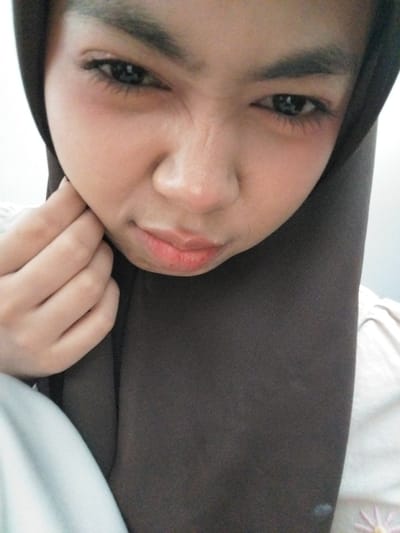 This is a close-up, portrait-style image of a young person wearing a dark brown hijab and lying down. The subject's face is tilted, with their eyes looking towards the viewer. Their expression appears somewhat wistful or pensive. The skin tone is light with a visible blush on the cheeks. The lips are slightly parted, revealing a hint of color, and the eye makeup includes mascara. The subject's left hand is visible, resting near their face, with fingers gently curled.

The background consists of soft, neutral fabrics, suggesting an indoor setting, possibly a bed or couch. The lighting is soft and diffused, indicating natural light, perhaps from a window, or a gently lit indoor environment. The overall mood is intimate and personal.

There is no visible text in the image. The focus is entirely on the subject's face and their implied emotional state. The image was taken in Tibojong, Indonesia.