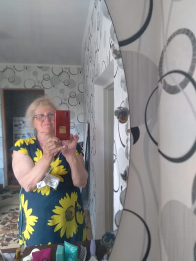 An older woman takes a mirror selfie indoors in Журавское сельское поселение, Ukraine. The image, originally rotated, shows the woman with blonde hair, wearing glasses, and a dark blue top adorned with prominent yellow sunflower patterns. She holds a red smartphone with a yellow sticker on its back, capturing her reflection. Her fingernails are painted with a light pink polish.
The setting appears to be a room with light-colored walls and a ceiling, both featuring a modern wallpaper design of various grey and black concentric circles and abstract patterns. In the reflection, to the right of the woman, a shelf or counter holds several personal items, including a hairbrush, small tubes or bottles (some blue, one pink), and a golden cosmetic item. A white power outlet or extension cord is visible near her right arm, with wires extending from it. The floor or a rug with a brown and beige patterned design is partially visible at the bottom right. The overall activity is a casual self-portrait taken within a residential interior. No specific time of day or visible text is discernible.