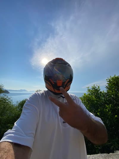 A selfie is taken by a person wearing a full-face orange and black motorcycle helmet, obscuring their face. The person is also wearing a white t-shirt and is making a peace sign with their right hand. The background shows a bright blue sky with scattered clouds and the sun shining intensely. Below the sky is a body of water, likely the sea, with distant islands or mountains visible on the horizon. Lush green foliage frames the right side of the image, and a stone ledge is visible in the foreground. The scene suggests an outdoor location, potentially a scenic overlook or a coastal area. The location is identified as Cam Lam, Vietnam.