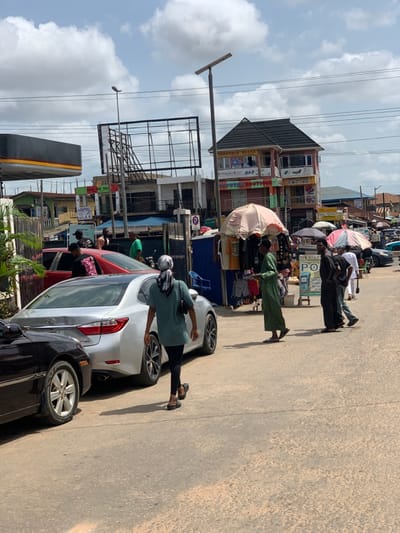 The image shows a street scene in Akure, Nigeria, at the coordinates 7.252 latitude and 5.198 longitude. There are several people walking on the street, some of whom appear to be shopping at various roadside stalls. A silver car is parked in the middle of the street, and a black car is parked on the left side of the street. The street is paved and dusty. In the background, there are several buildings, including a two-story building with a sign that reads "New Boutique." There is also a large billboard that is blank. The sky is partly cloudy. The people in the foreground are wearing various clothing. Some of them are carrying bags or purses. One person is wearing a headscarf. The environment is busy and chaotic, with traffic, pedestrians, and vendors all vying for space.