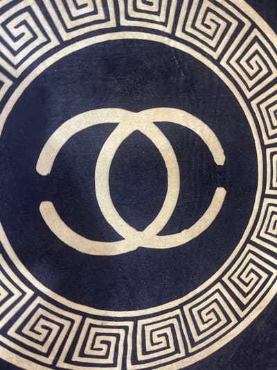 The image displays a close-up view of a dark circular pattern, likely on a fabric or rug. The pattern features a prominent golden interlocking "CC" logo in the center, reminiscent of the Chanel brand. Encircling the logo is a decorative border composed of a Greek key or meander pattern, also in golden lines against the dark background. The texture of the dark material appears to be slightly velvety or fuzzy. The image is an artistic or decorative close-up, with no environmental context provided. There are no people, actions, or specific city locations visible in the image.