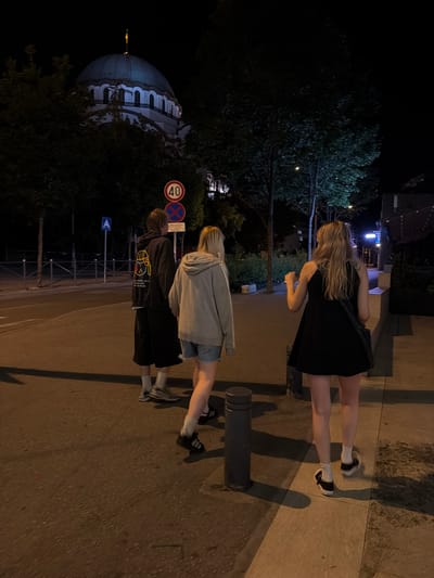 The image depicts a nighttime scene on a street in Vracar Urban Municipality, Serbia, with a prominent domed building in the background. Three individuals are walking away from the camera on a sidewalk adjacent to a street. The person closest to the camera on the right is wearing a black dress and white socks with black shoes. To their left, another person is wearing a grey hoodie and denim shorts. The furthest person, on the left, is wearing a dark hoodie with a graphic print on the back, black shorts, and white socks with grey sneakers. A traffic sign indicating a 40 km/h speed limit and another sign with a red circle and a diagonal bar are visible on the left side of the street, along with a blue directional sign. Trees line the street, and streetlights are casting shadows. The overall atmosphere is dark and urban.