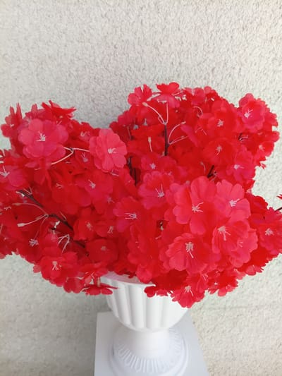 A bouquet of vibrant red artificial flowers is displayed in a white urn-shaped vase. The vase is placed on a white pedestal. The background is a textured, light-colored wall. The image is a close-up shot of the flowers and vase, with the wall filling the upper portion of the frame. There are no people or animals present in the image. The location in the city of Osh, Kyrgyzstan is not discernible from the visual information provided in the image.