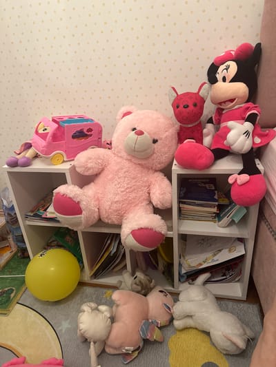 The image shows a white shelving unit filled with stuffed animals and books, resting on a patterned rug and in front of a polka-dot wall. The stuffed animals include Minnie Mouse, Mickey Mouse, a pink teddy bear, a red rabbit, a white cat, a pink pony with wings, and a smaller gray teddy bear. A yellow balloon is also visible near the bottom of the shelves. A pink plastic toy car is near the teddy bear. There is no evidence to indicate the location of the photo is in Esenyurt, Turkey.