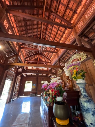 Intricate wooden beams create a geometric ceiling pattern above a room decorated with religious iconography. Statues, floral arrangements, and decorative plates adorn a surface, while windows provide natural light. The architecture and decorations suggest a temple or place of worship, showcasing detailed craftsmanship and spiritual significance. The structure emphasizes height and ornate design.