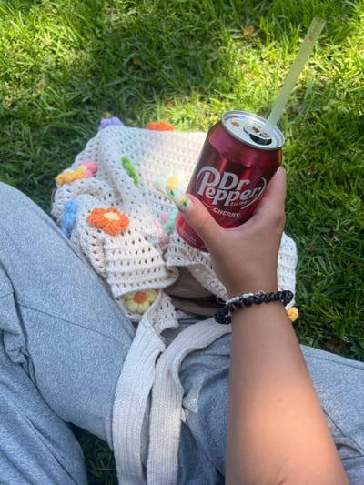 The image depicts a close-up, overhead view of a person's hand holding a can of Dr Pepper Cherry with a straw, resting on a crocheted garment with flower appliques. The person is wearing grey sweatpants and a bracelet with black and pearl beads. The scene is set on a grassy lawn, likely in Kiryat Bialik, Israel, suggesting an outdoor leisure activity. The lighting indicates a sunny day.